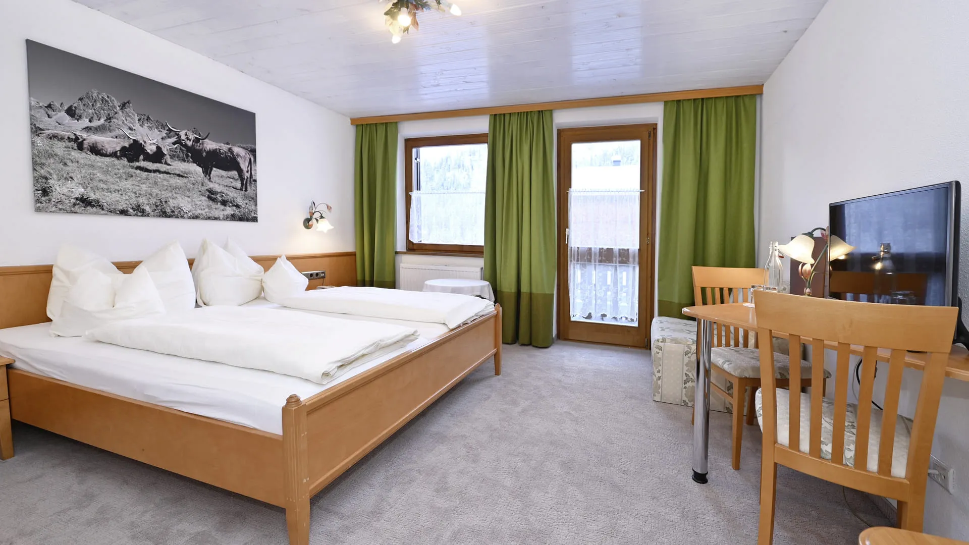Helles Schlafzimmer mit Doppelbett, Schreibtisch und Fenster mit grünen Vorhängen.